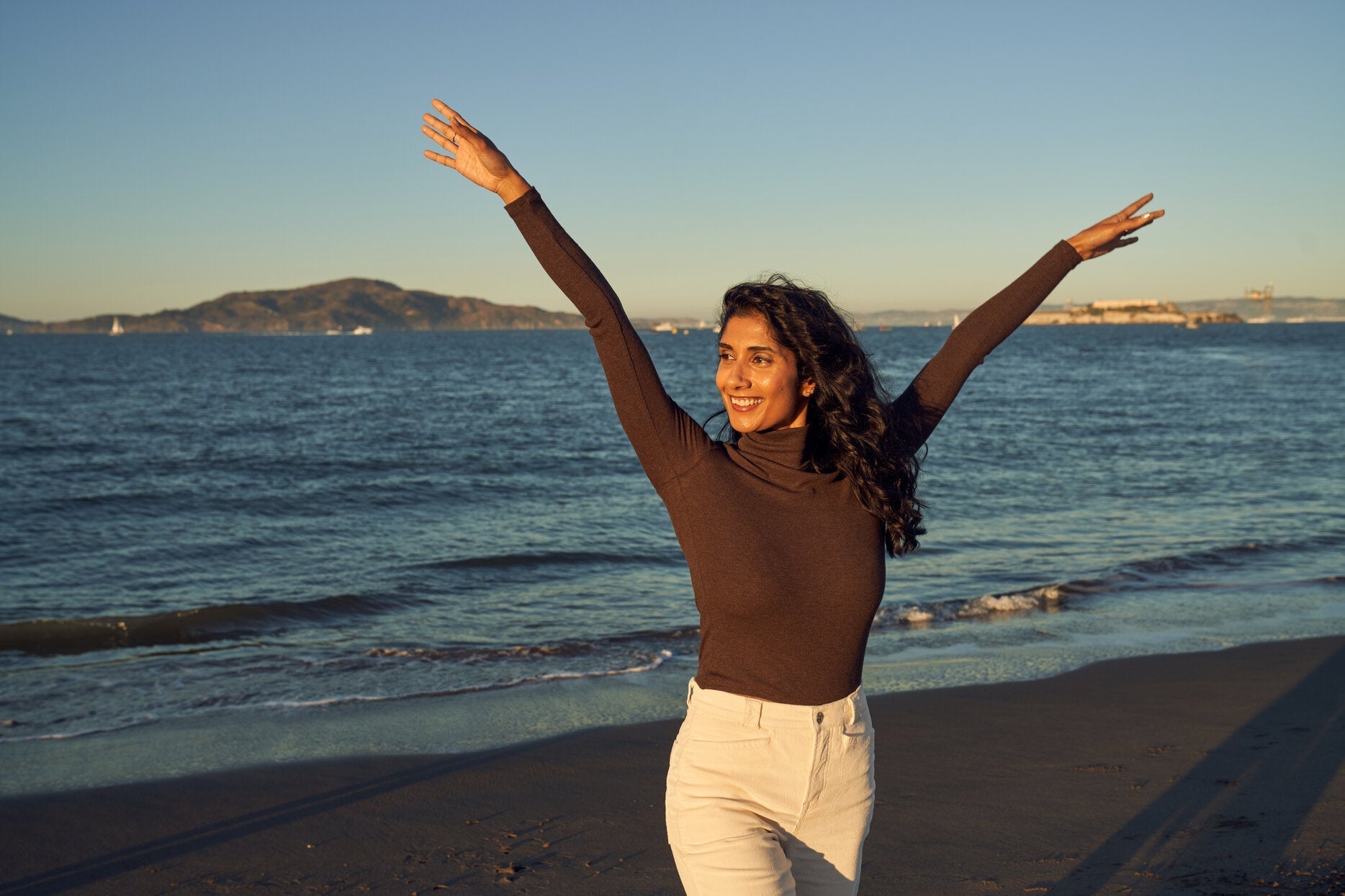 A woman smiles with her arms raised joyfully while standing on a beach at sunset. She is wearing a long-sleeved brown UPF-rated top and light-colored pants.