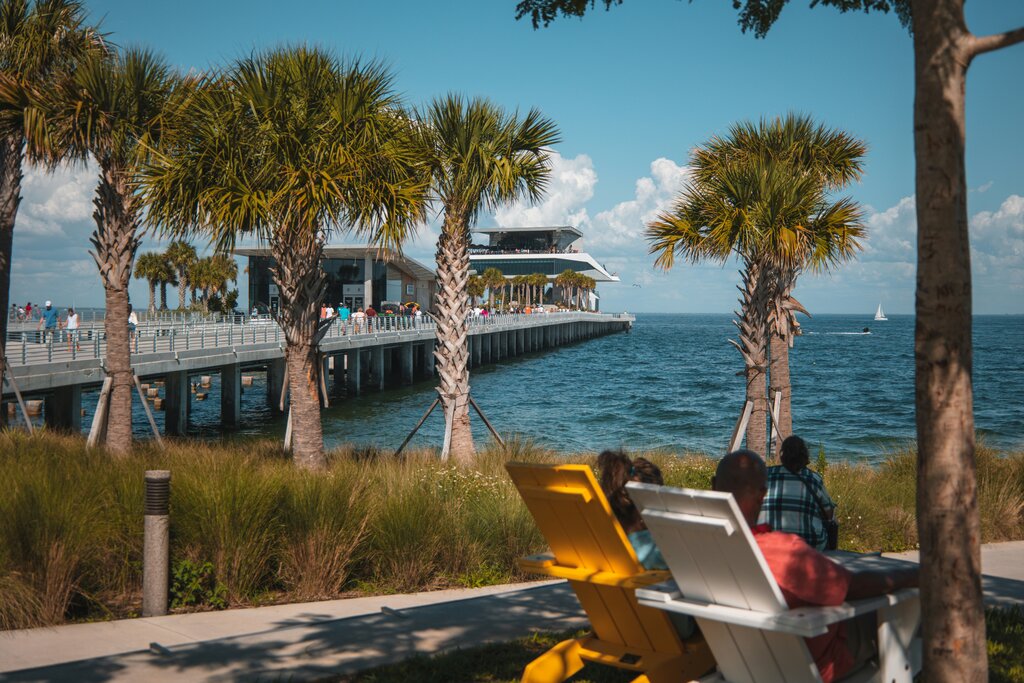 People relaxing in colorful chairs along the waterfront in St. Pete, Florida, with a scenic view of The St. Pete Pier surrounded by palm trees and calm blue waters.