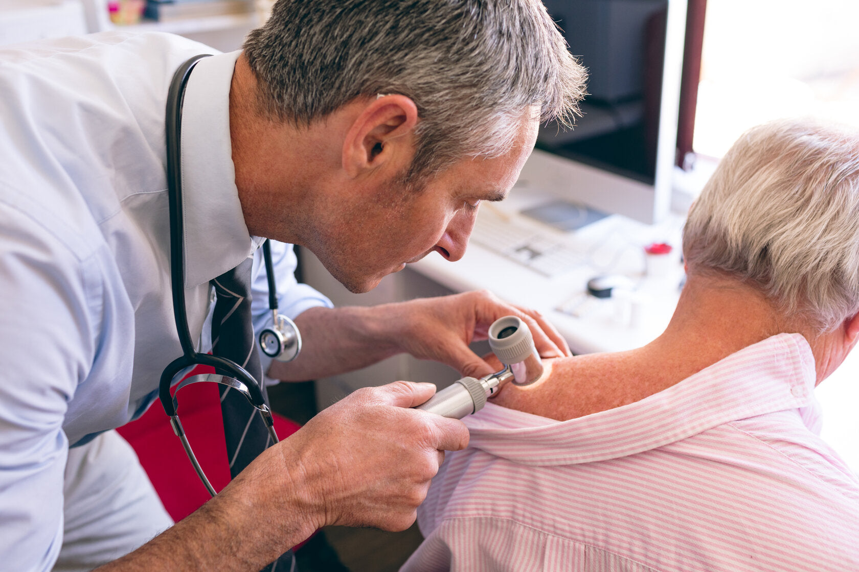 Doctor examining an older patient's shoulder with a dermatoscope, focusing closely on skin health and potential signs of skin cancer.