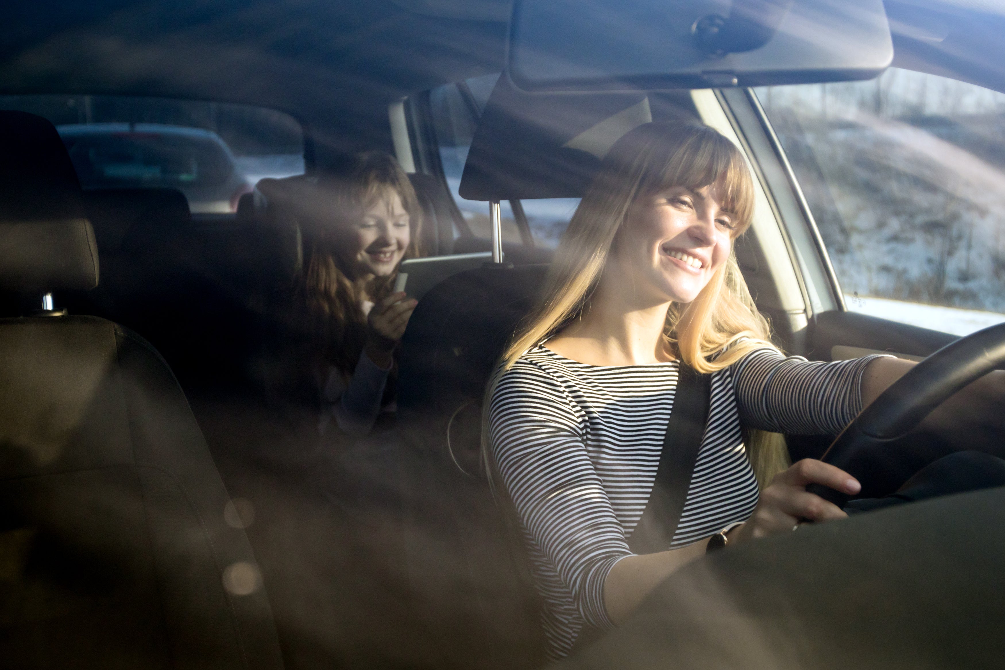 A smiling woman driving with sun rays streaming through the car windows, highlighting sun exposure inside the vehicle, while a young girl sits in the back seat.