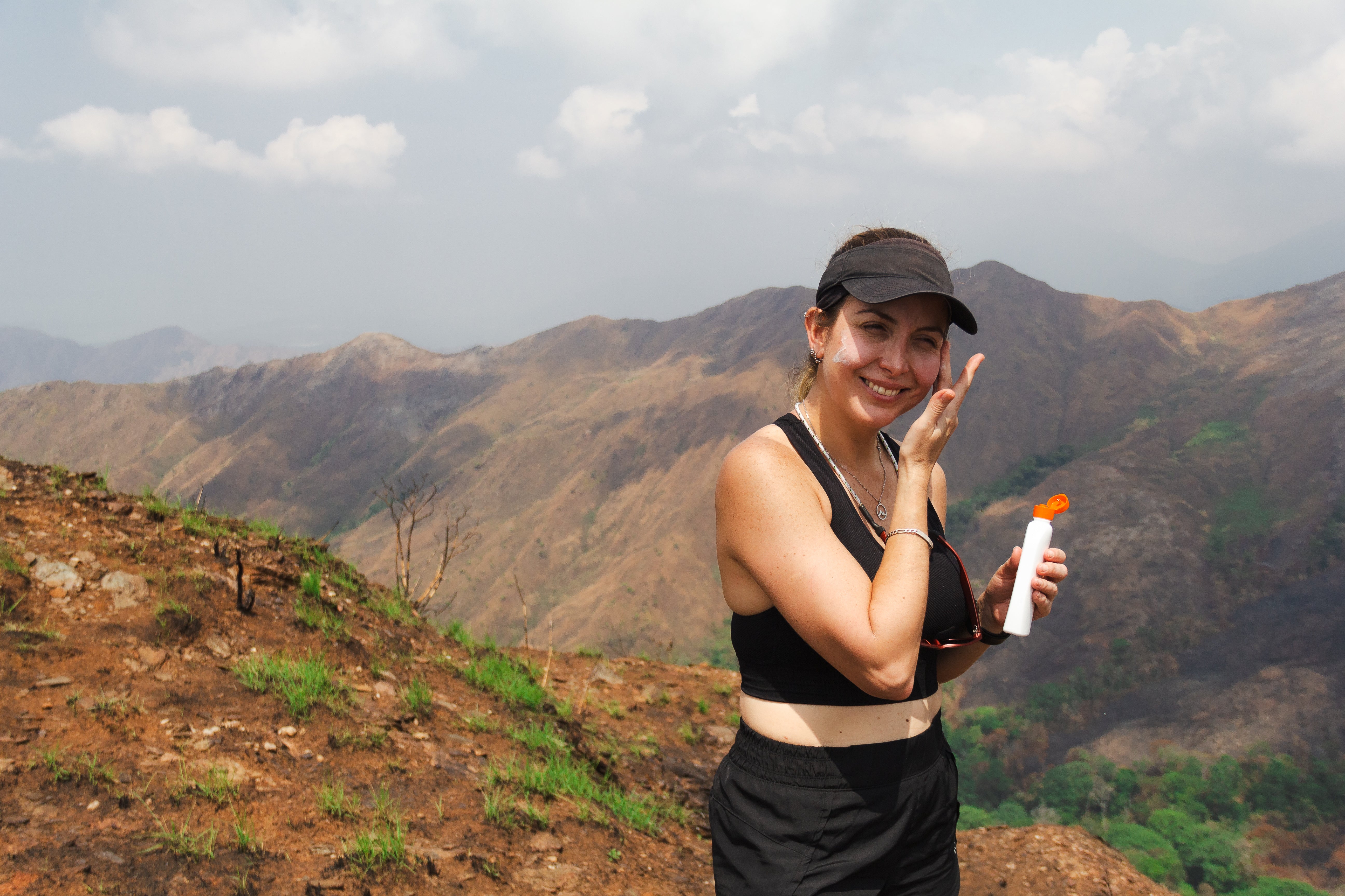 A woman applying sunscreen on a mountain trail, wearing a black visor and athletic gear with scenic mountains in the background.