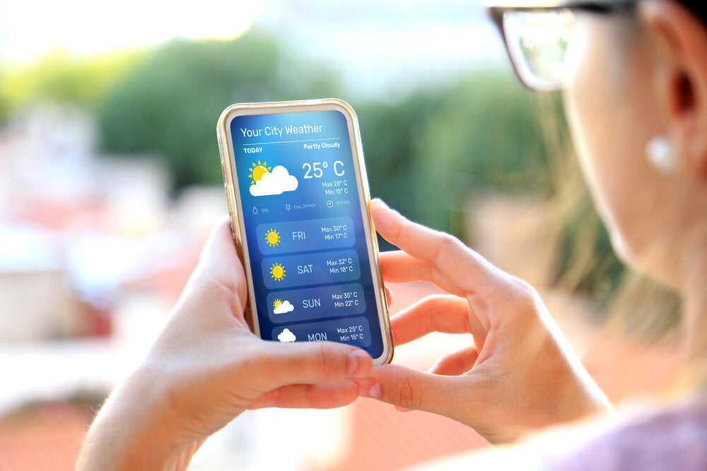 Close-up of a woman holding a smartphone displaying a weather app, showing the weekly forecast and current temperature of 25°C under partly cloudy skies.