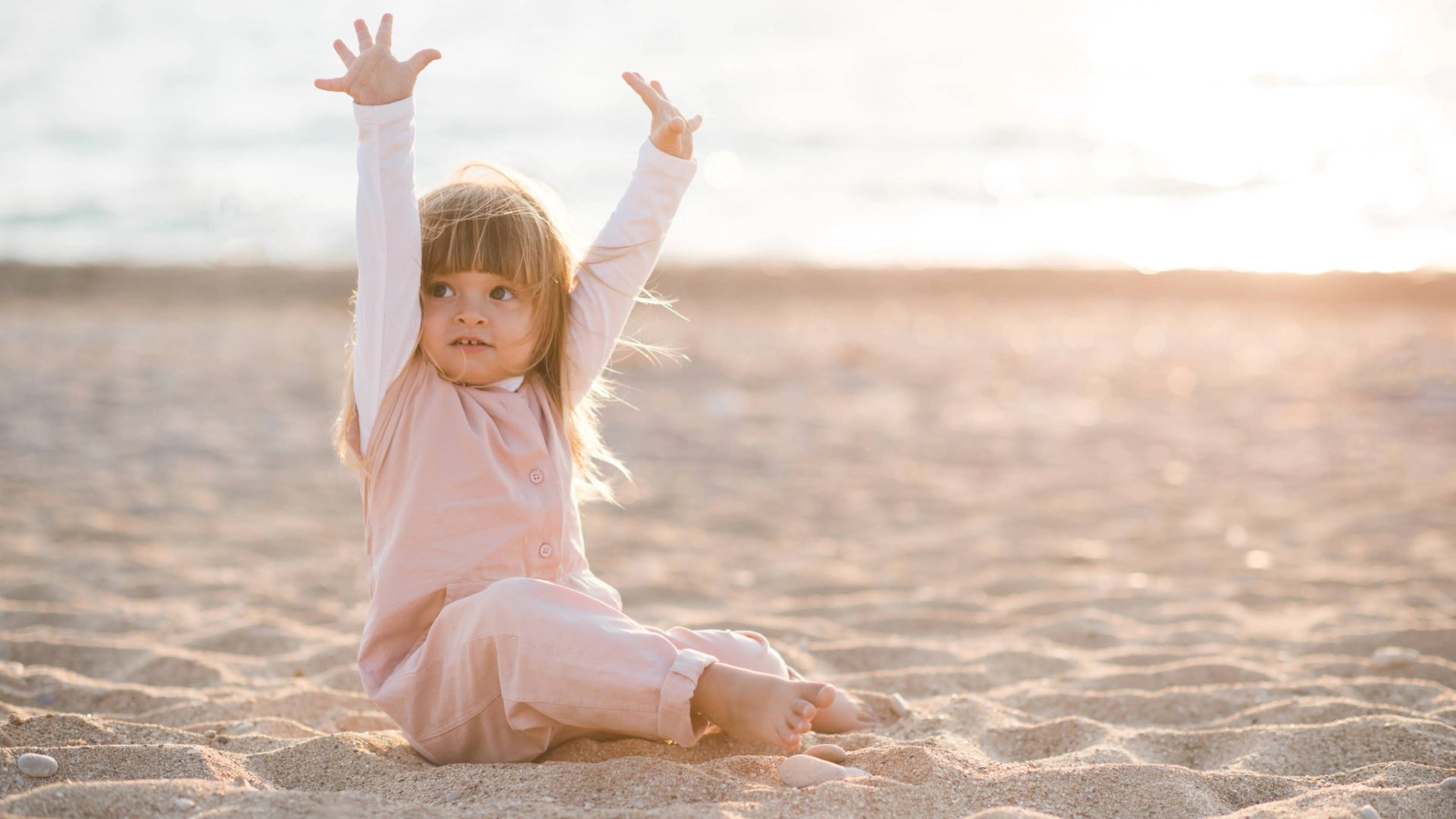 Young child sitting on the beach at sunset, raising arms playfully while wearing a long-sleeve UPF-rated outfit