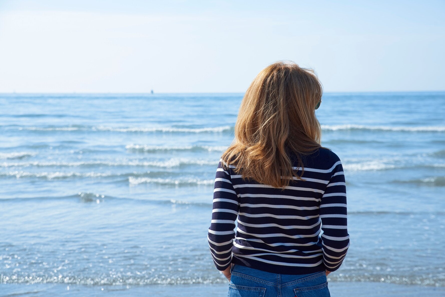 Woman standing on the beach, facing the ocean on a sunny day, wearing a striped long-sleeve UPF-rated shirt