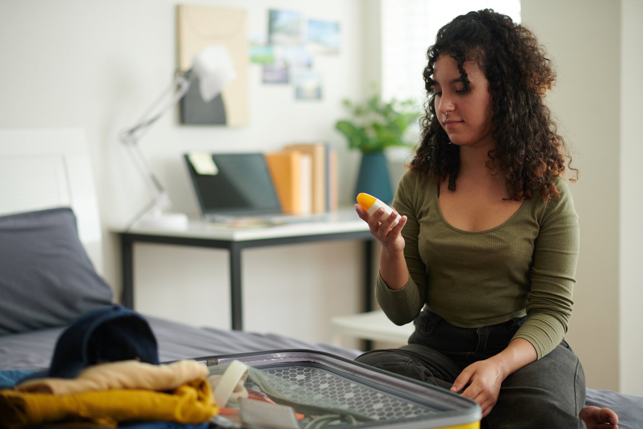 A young woman sitting on her bed, holding a bottle of sunscreen while preparing to pack her suitcase.