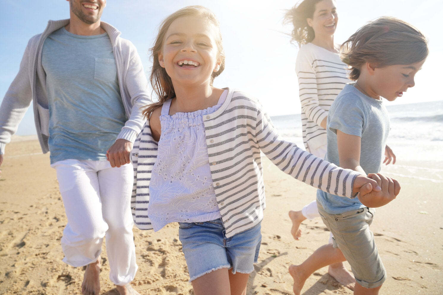 A joyful family runs on the beach, with the children and parents wearing light, UPF-rated clothing, enjoying the sunny day safely under the sun's rays.