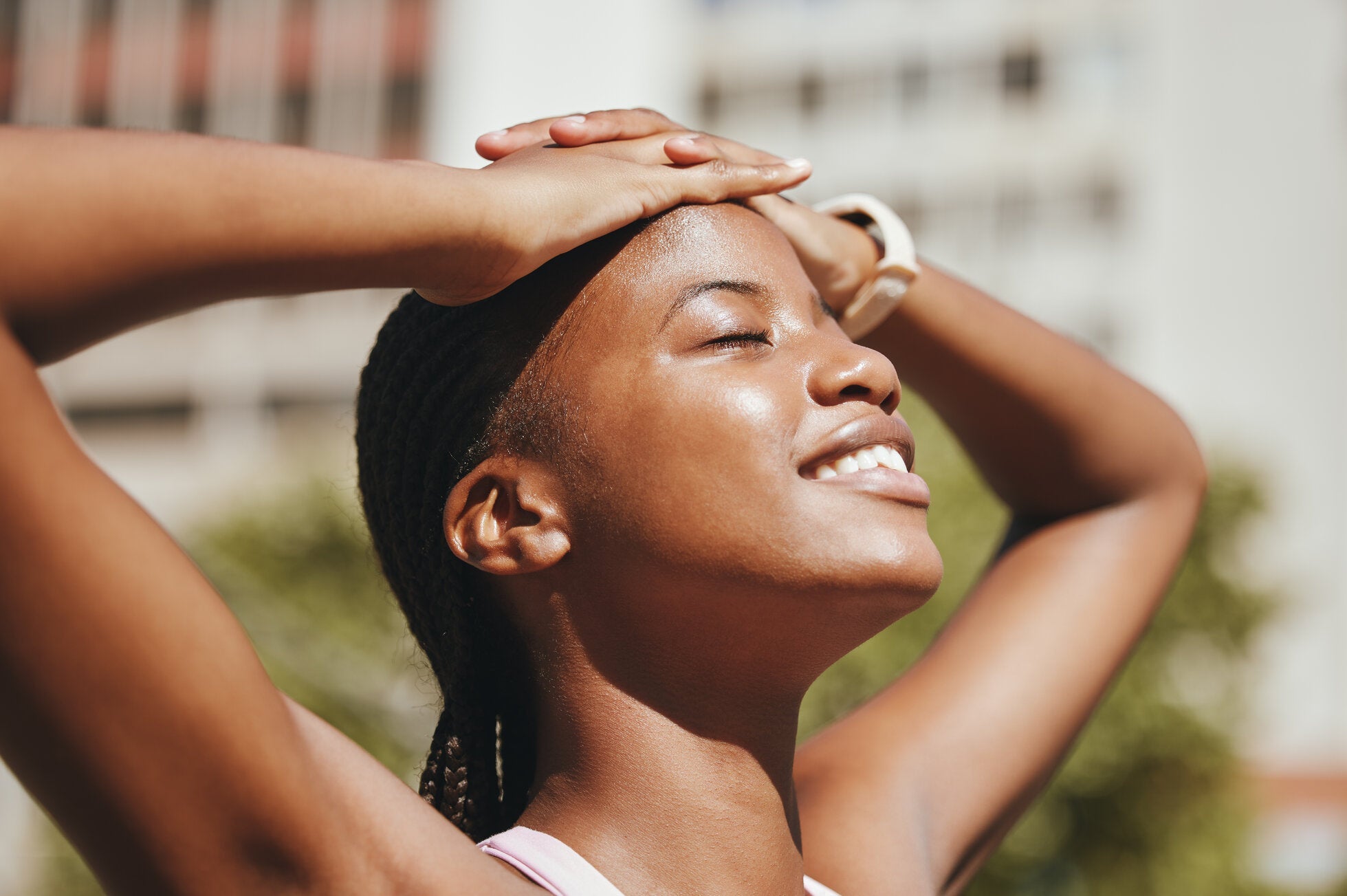 A woman smiling with her eyes closed, enjoying the sunlight, with her hands resting on her head.