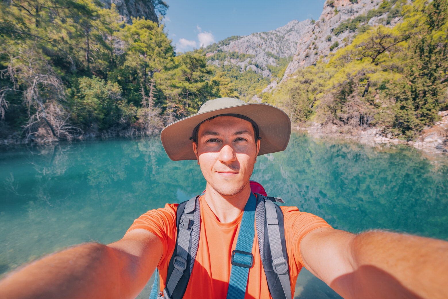 A man taking a selfie while hiking, wearing a wide-brimmed UPF-rated hat and backpack, with a turquoise lake and forested mountains in the background.