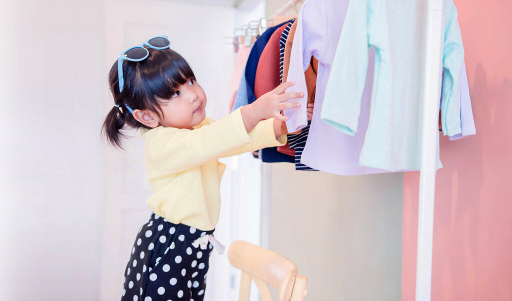 A young girl wearing sunglasses and a stylish outfit reaches for a long-sleeve UPF shirt from a clothing rack, examining multiple colors of sun-protective clothing for her wardrobe.