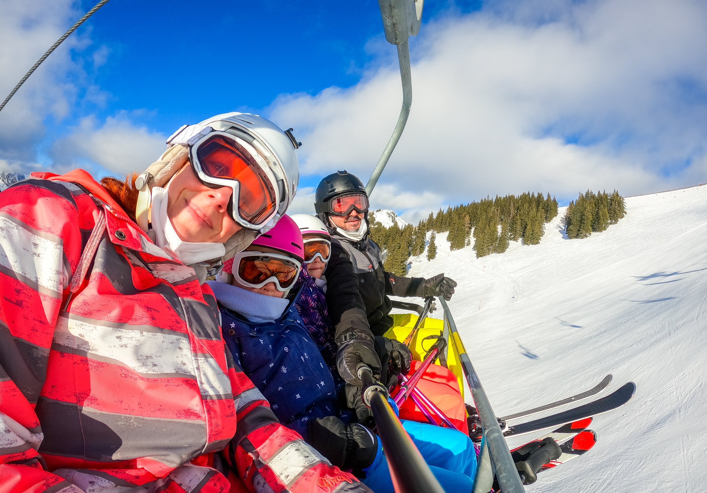 A family of skiers takes a selfie on a ski lift, surrounded by snowy mountains and bright sunshine.