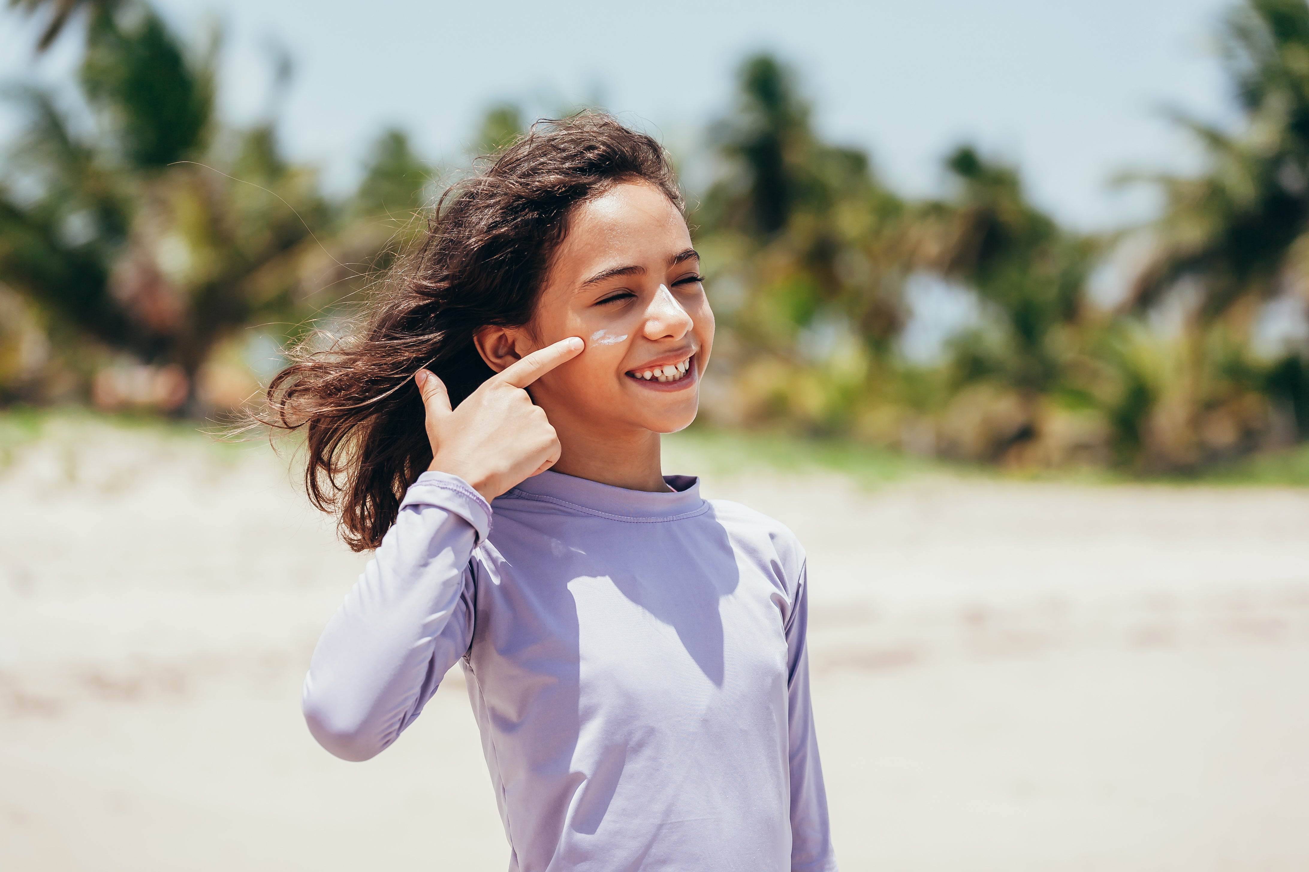 A young girl applying sunscreen on her face at the beach, wearing a long-sleeve UPF shirt, with palm trees in the background.