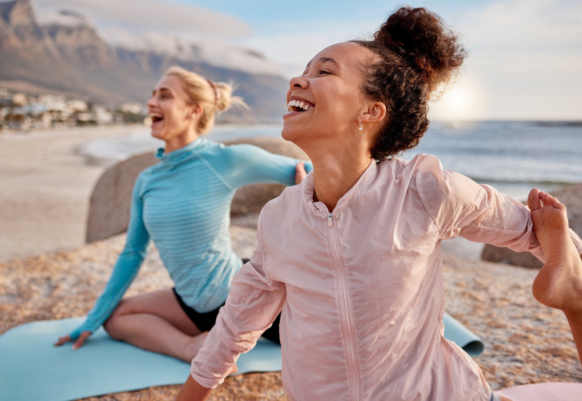 Two women smiling and stretching during a yoga session on a beach, with mountains and the ocean in the background.