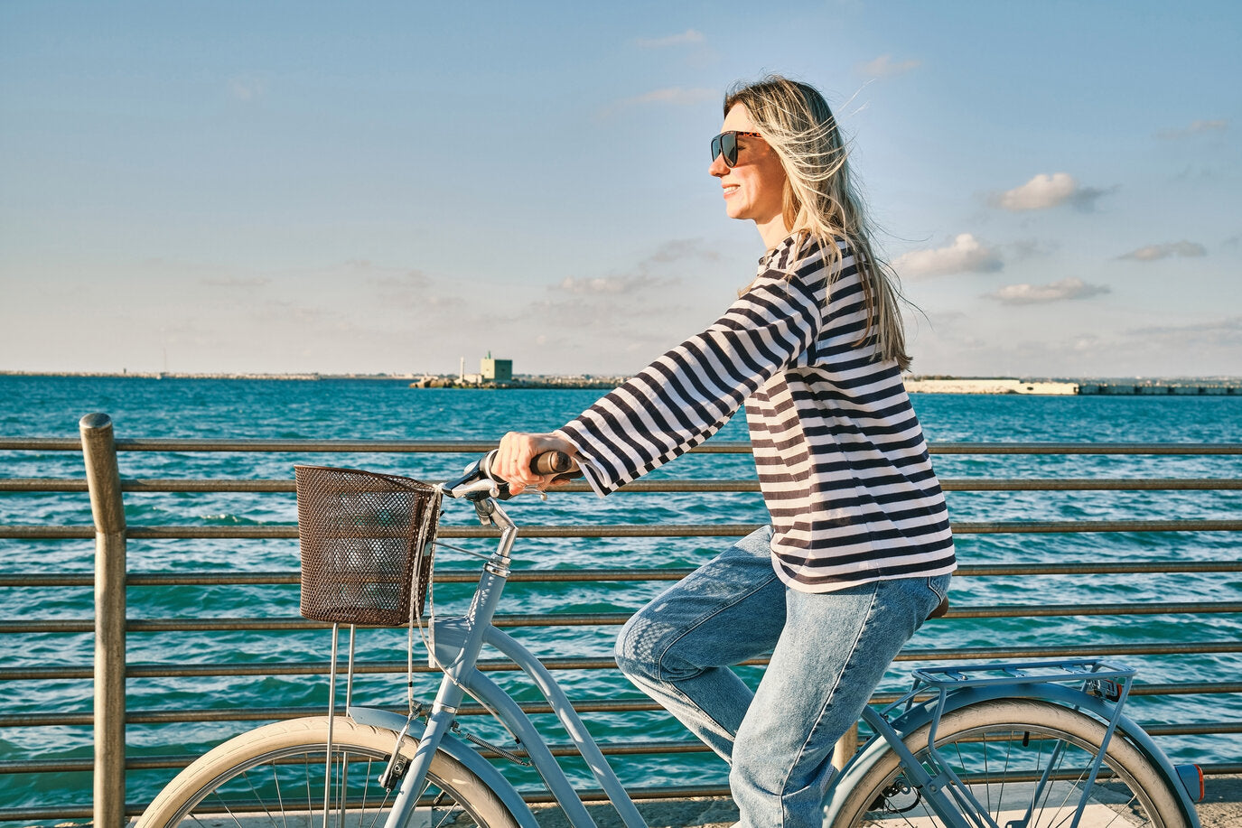 A woman riding a bicycle along the waterfront on a sunny day, wearing sunglasses and a striped long-sleeved UPF-rated shirt.