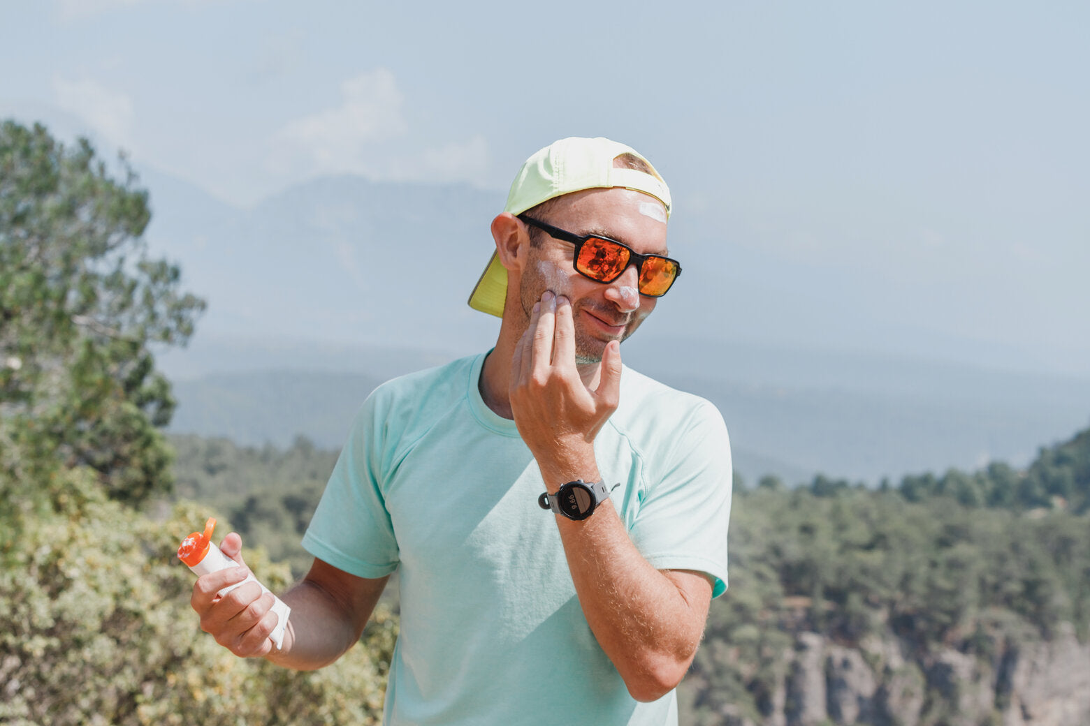 A man in a light blue t-shirt and sunglasses is applying sunscreen to his face while standing outdoors in a sunny, mountainous area. He wears a backward cap and holds a sunscreen bottle in his other hand, preparing for sun exposure. 