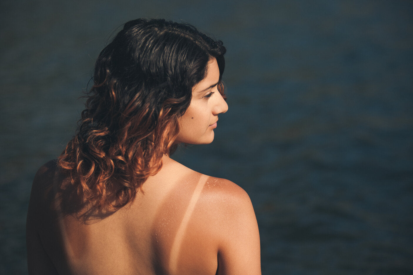 Young woman with sun-kissed skin and a visible tan line on her shoulder, standing outdoors near the water.