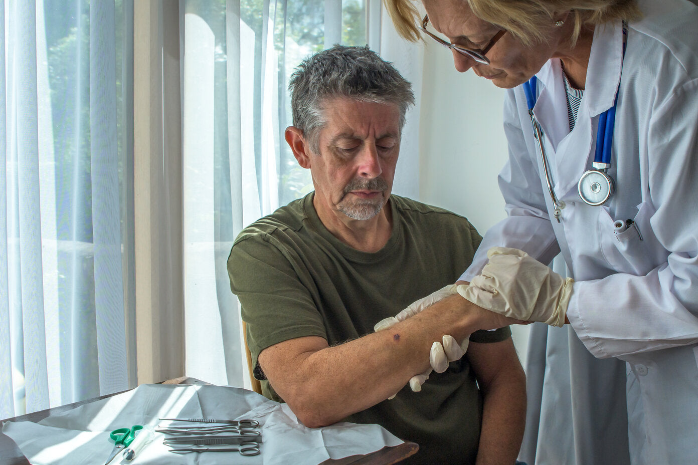 Older man sitting while a doctor wearing a white coat and gloves examines a spot on his arm, with medical instruments laid out on a table nearby.