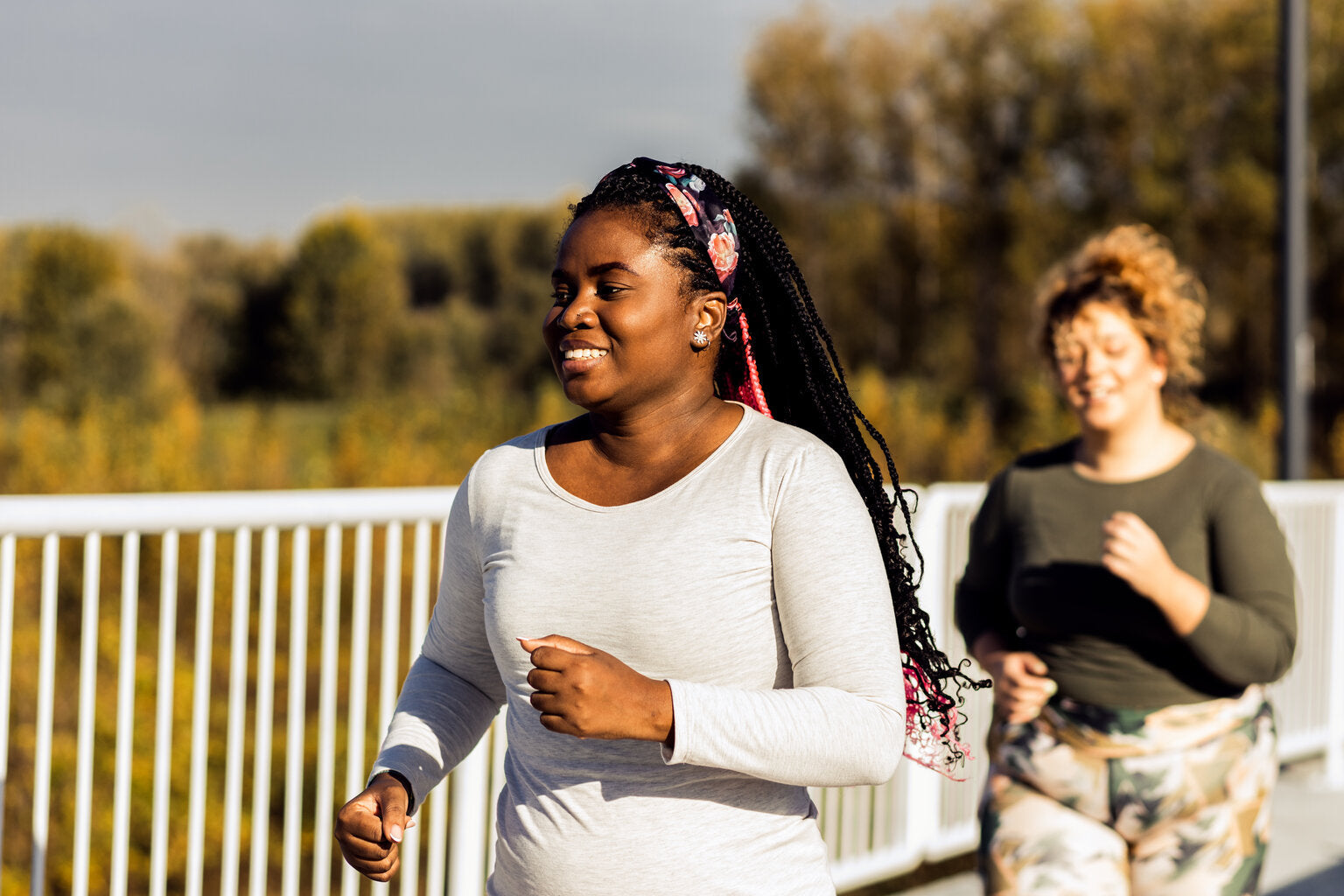 Two women jogging outdoors on a sunny day, smiling and enjoying their run. The woman in the foreground is wearing a long-sleeved light gray shirt and a colorful headband, with braided hair. Trees with autumn colors can be seen in the background.