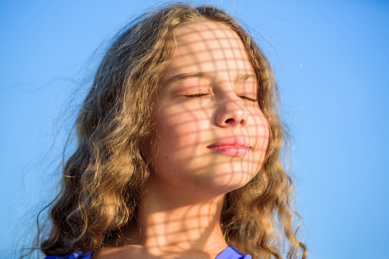 Young girl standing in the sun with eyes closed, shadow pattern on her face, enjoying a sunny day outdoors