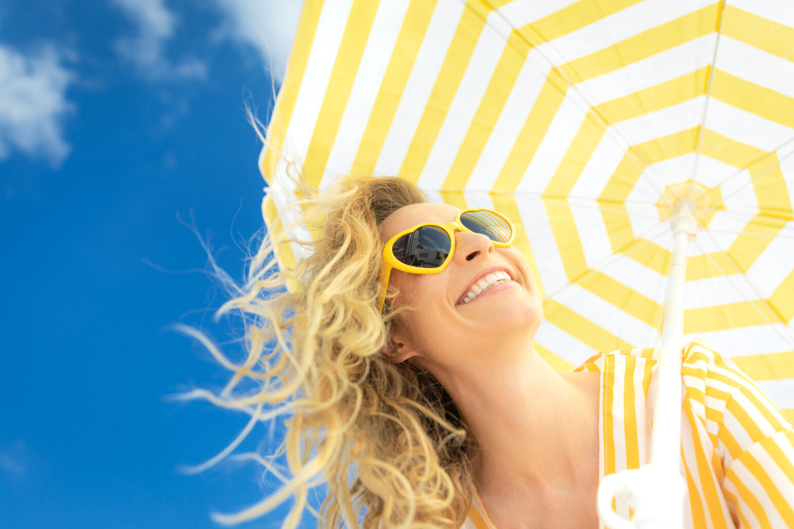 Smiling woman wearing yellow sunglasses under a yellow and white striped UVE-rated umbrella, enjoying a sunny day with a clear blue sky in the background.