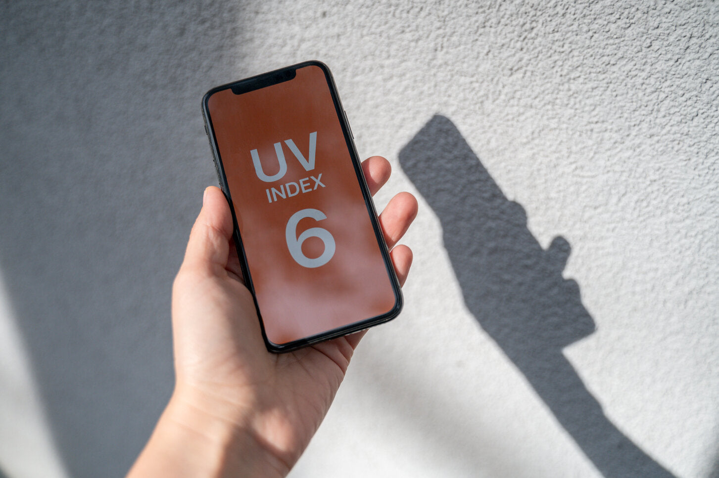 Person holding a smartphone displaying UV index 6, with sunlight casting a shadow on the wall