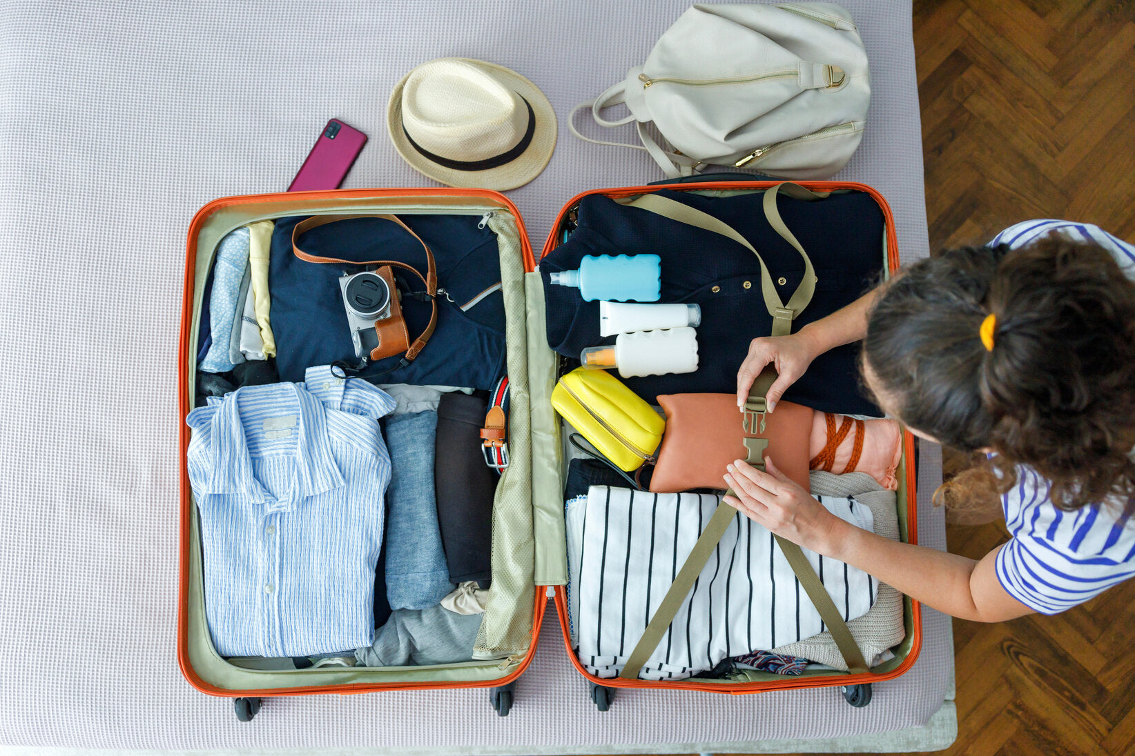 Overhead view of a woman packing a suitcase for a vacation. The suitcase contains neatly folded clothing, a hat, a camera, and travel-sized bottles, including sunscreen, suggesting preparation for a trip.