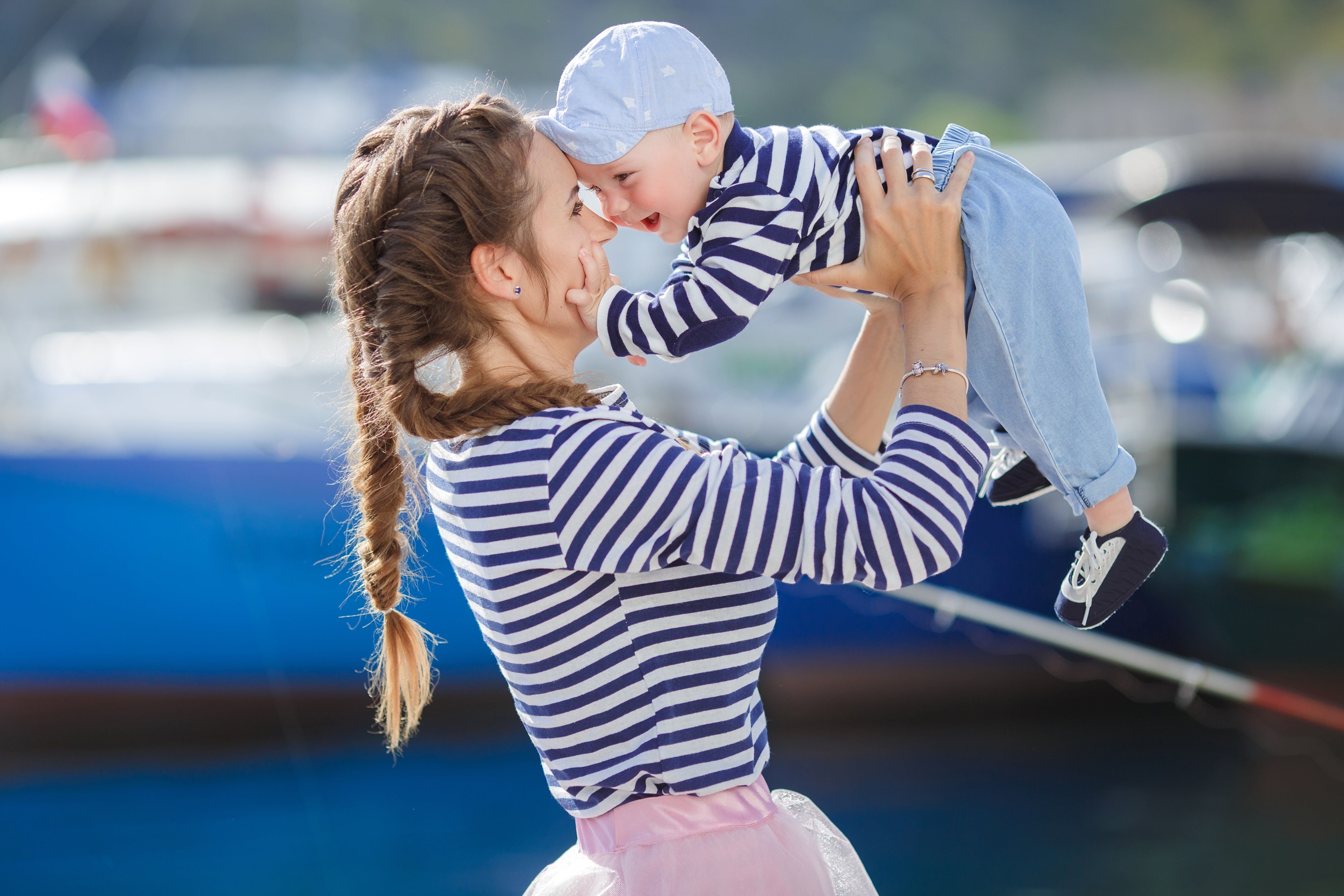 A mother lifting her baby in the air, both wearing matching striped outfits, with a marina in the background.