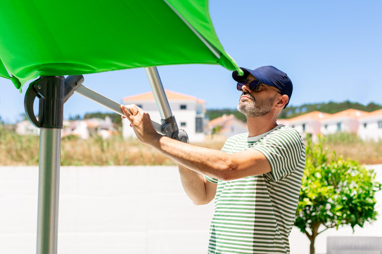 Man adjusting a bright green UVE-rated sun umbrella outdoors, wearing a striped shirt, sunglasses, and a hat to stay protected from the sun.