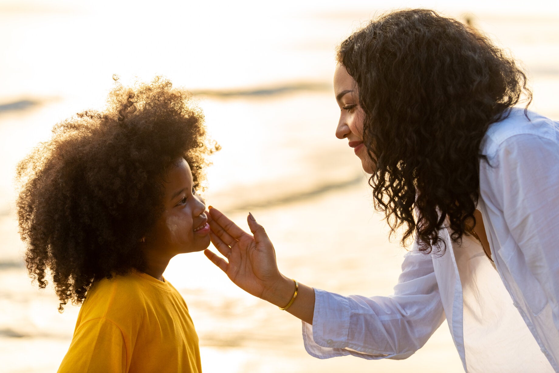 A mother gently applying sunscreen to her young child's face while at the beach. The child, wearing a yellow shirt, smiles as the mother, dressed in a white shirt, carefully ensures sun protection.