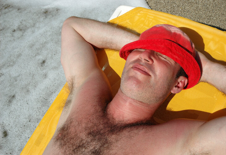 Man relaxing on a yellow lounge chair wearing a red hat, with visible sunburn on his chest, enjoying a sunny day outdoors despite sun damage.