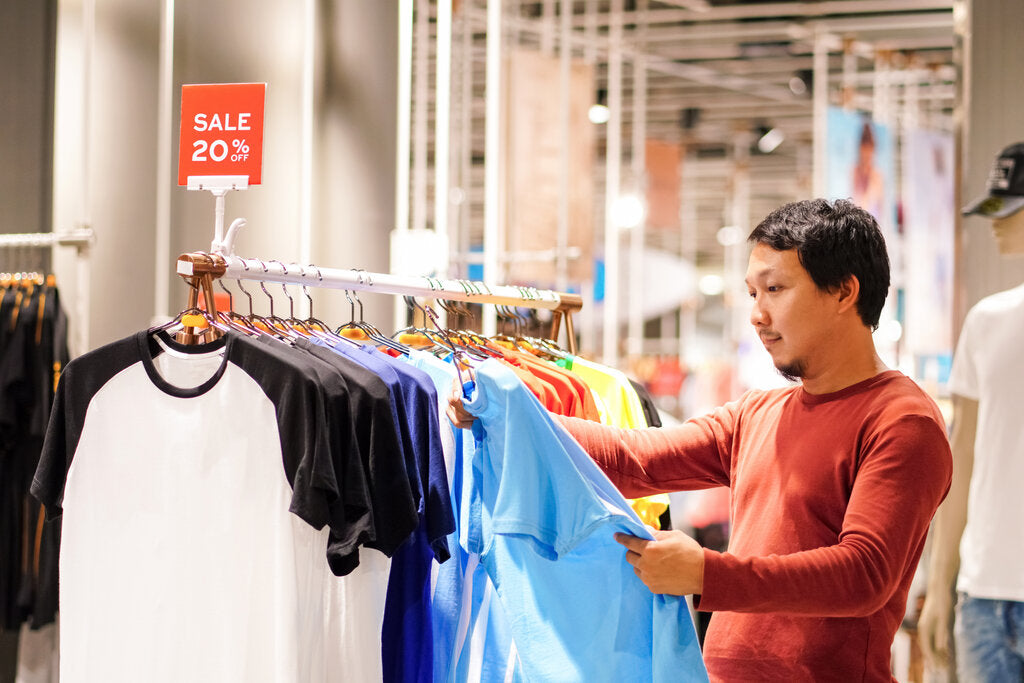 Man shopping for UPF shirts on a rack, carefully inspecting the lightweight fabric. A sign above highlights a 20% sale, promoting sun-protective clothing options.