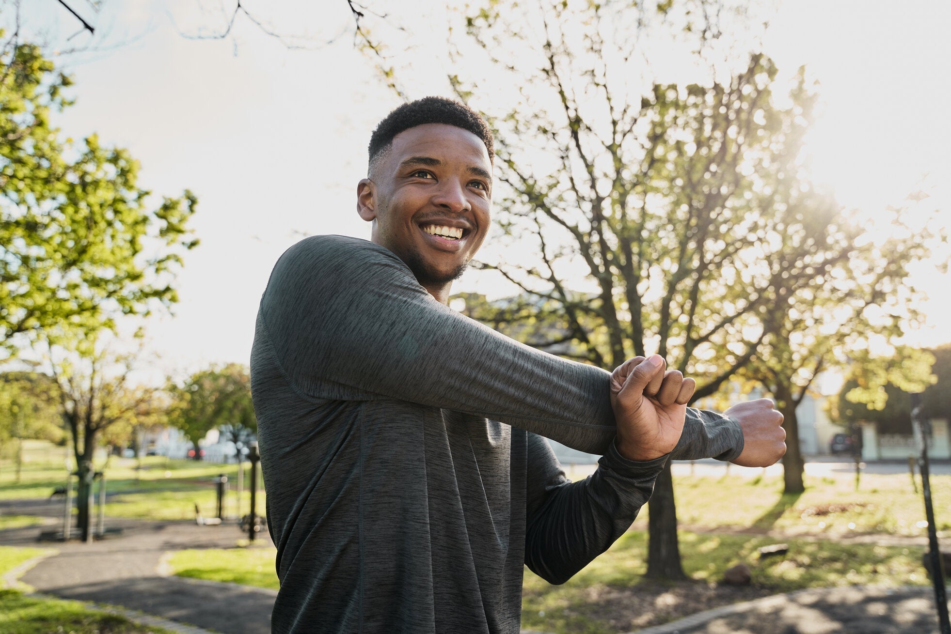 Man smiling as he stretches his arm outdoors in a sunny park, surrounded by green trees and warm sunlight filtering through.