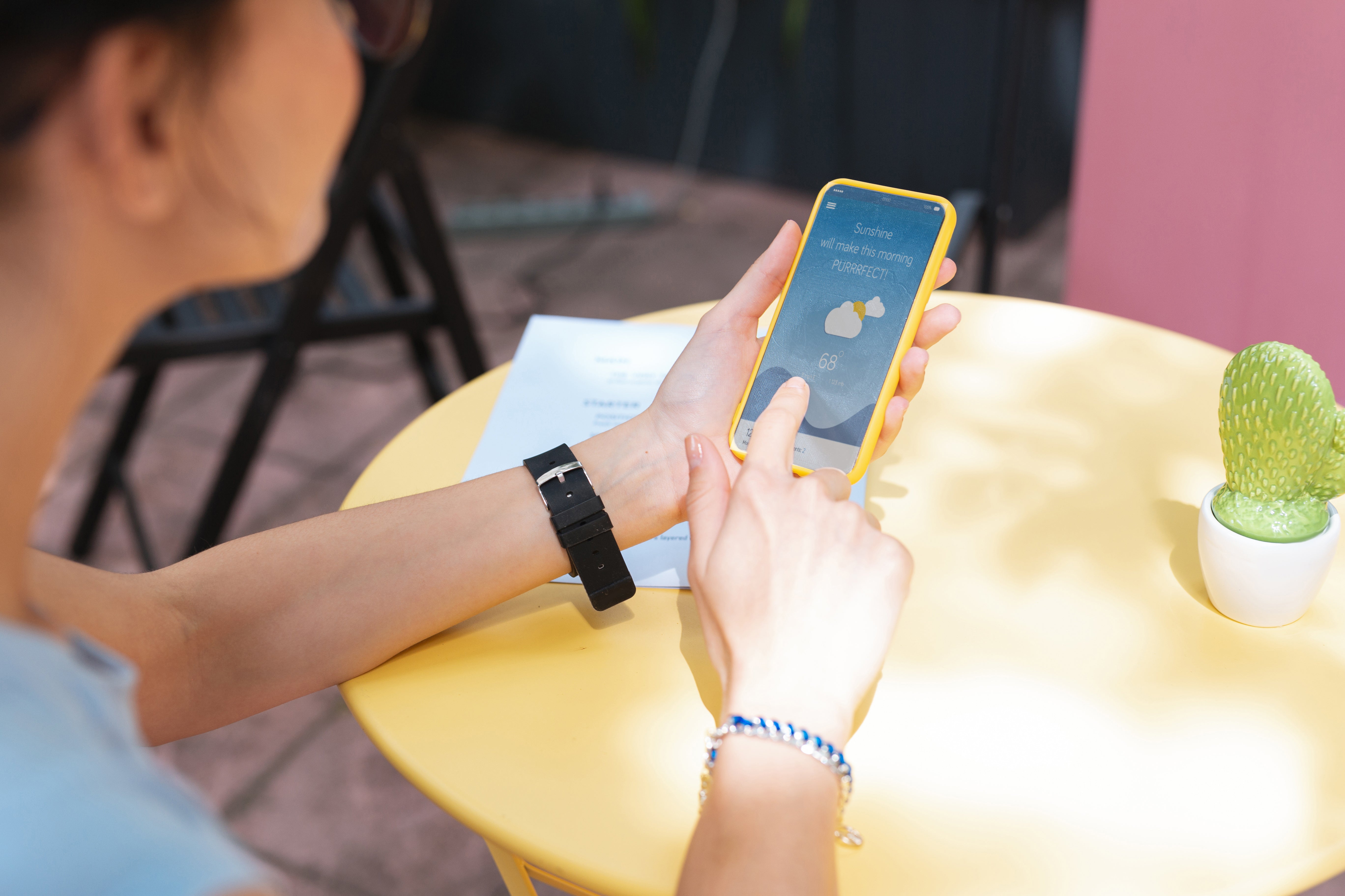 A person checking the weather and UV index on a smartphone while sitting at a yellow table outdoors.