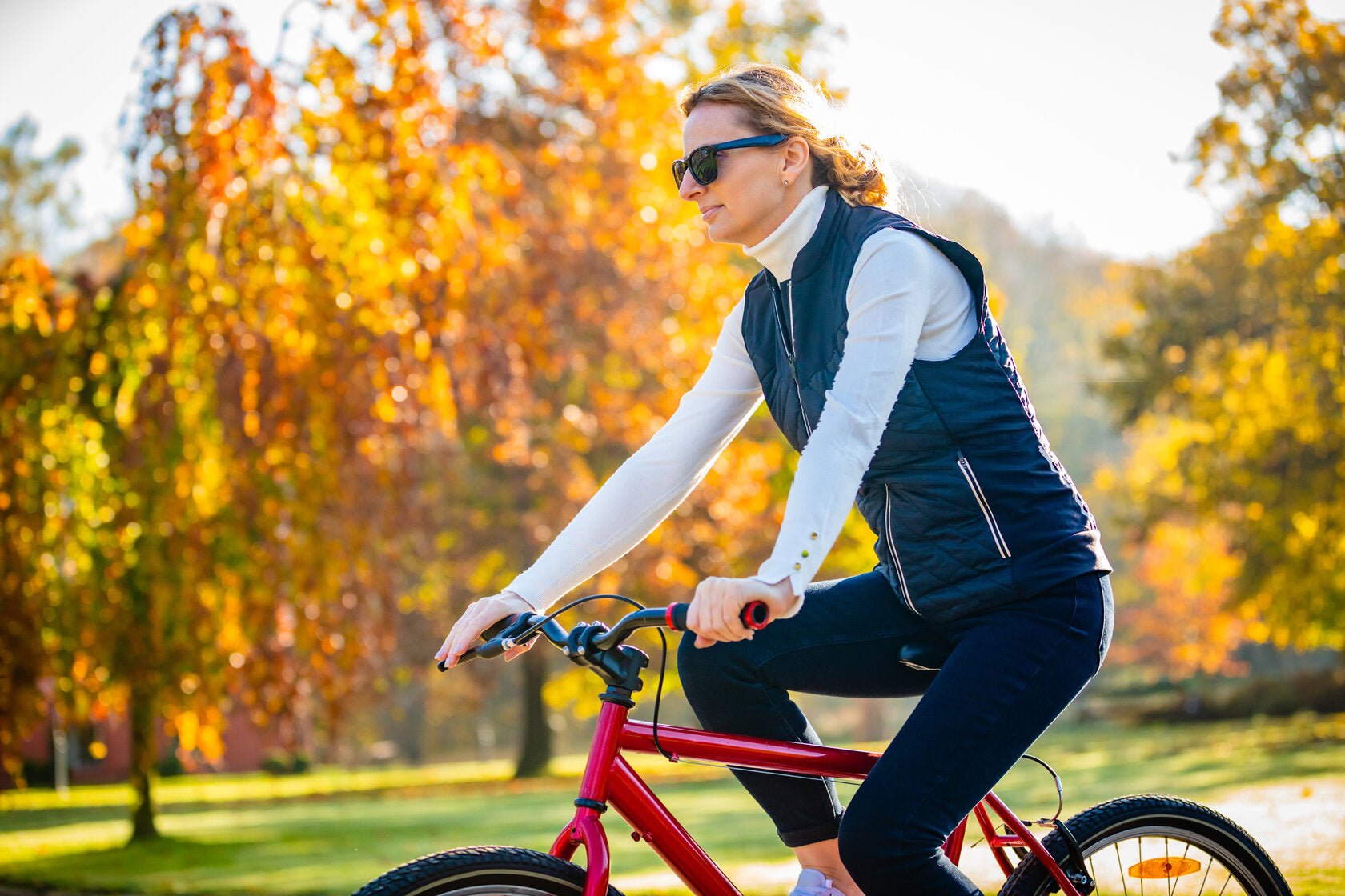 A woman riding a red bicycle on a sunny fall day, wearing a black vest, long-sleeved shirt, and sunglasses. The background features vibrant autumn trees with golden leaves, creating a scenic and peaceful outdoor setting.