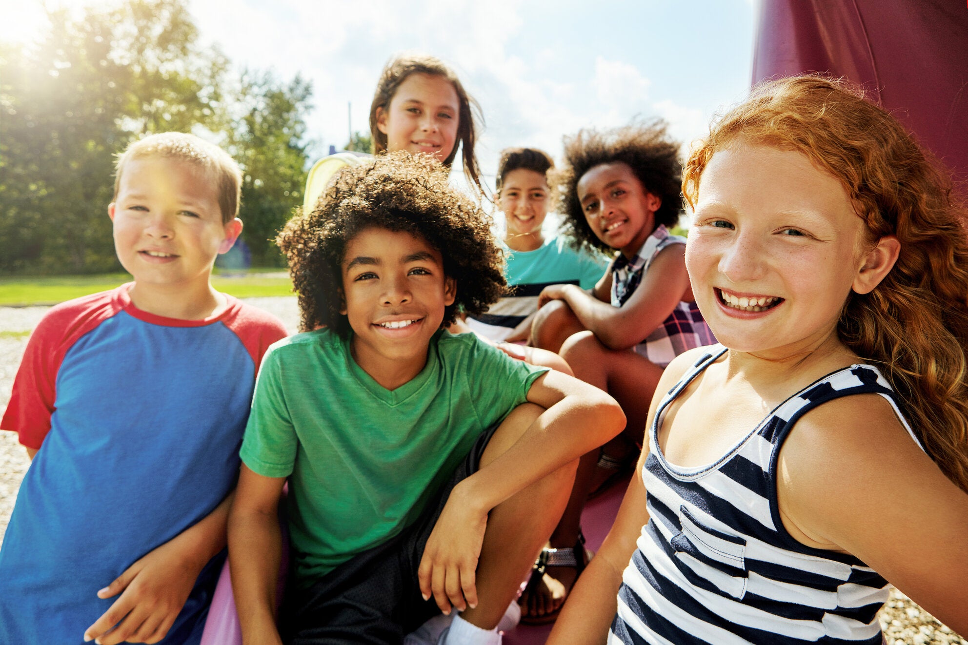 A diverse group of smiling children sits together outdoors on a sunny day, enjoying each other’s company on a playground.