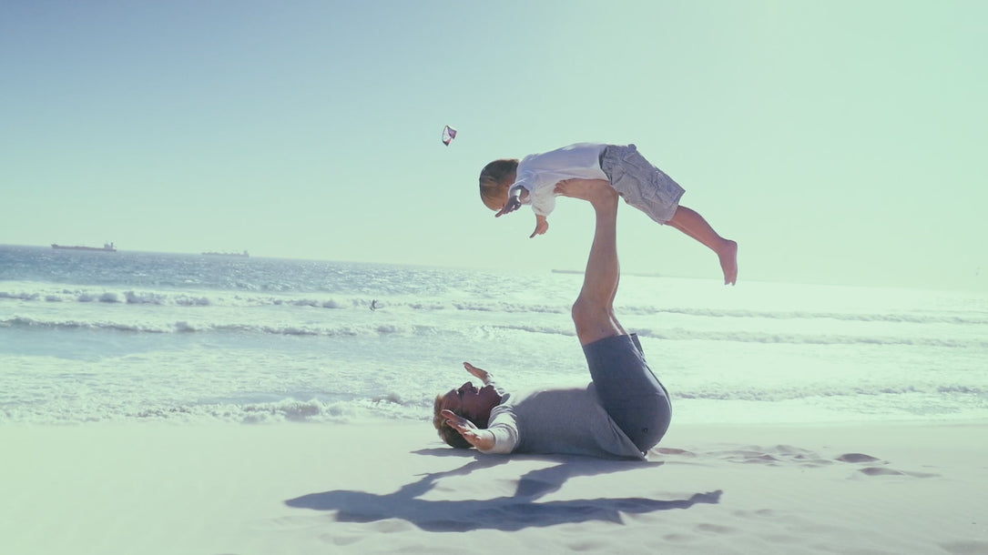 Adult balancing a child in an airplane pose on a sunny beach, with ocean waves and a kite in the background