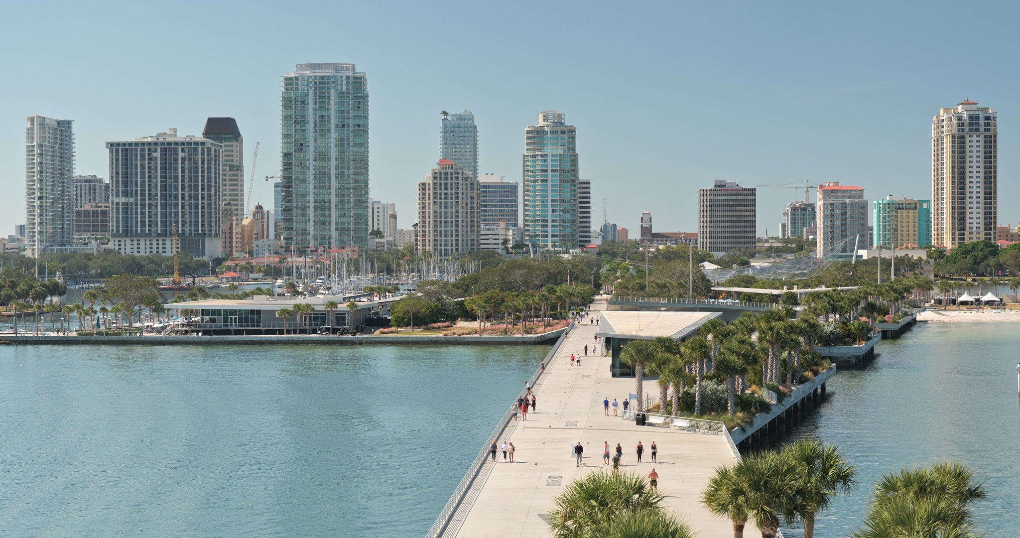 Scenic view of the St Petersburg skyline with tall buildings, waterfront, marina, and the St Pete Pier lined with palm trees and full of pedestrians on a sunny day