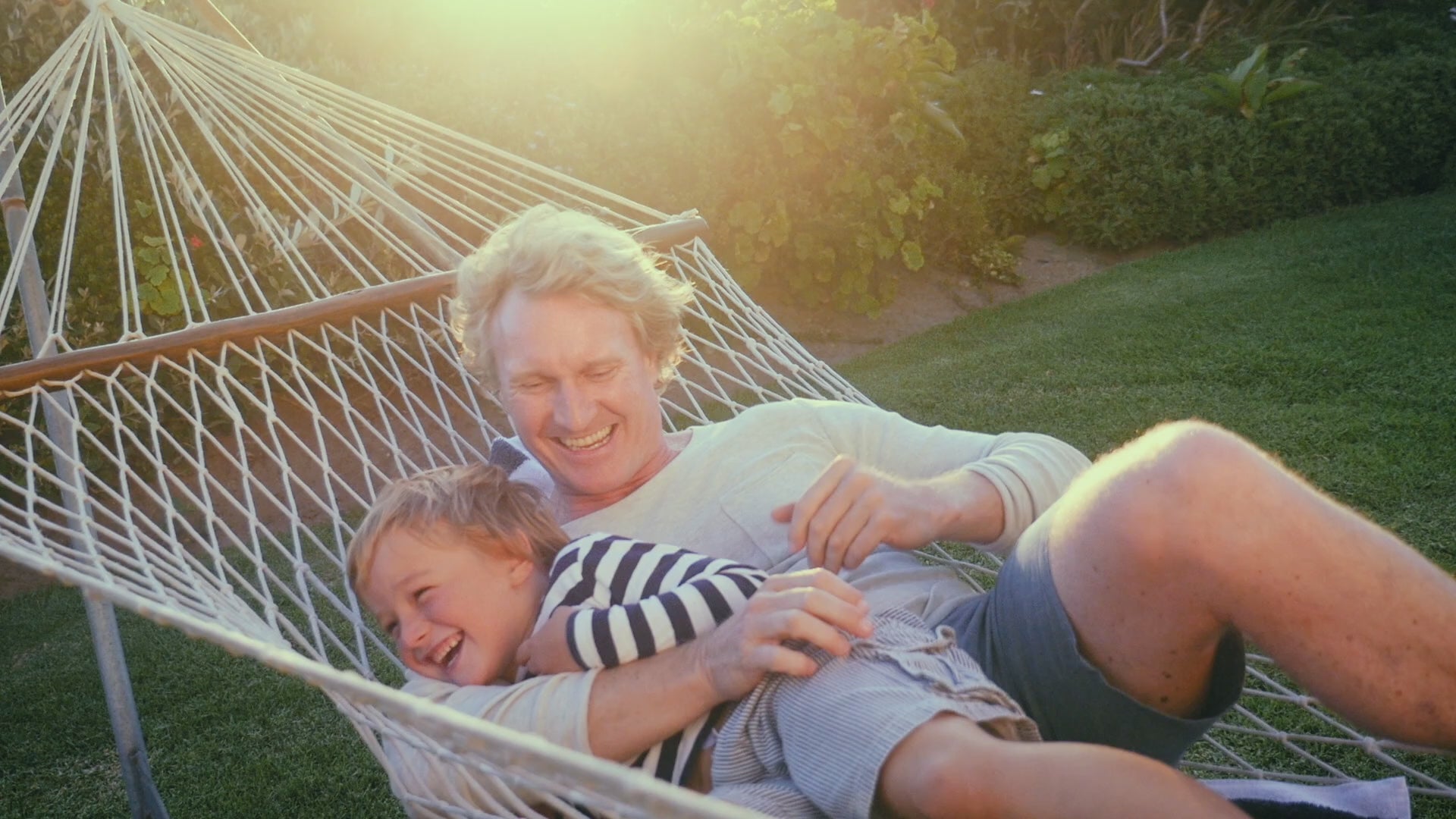 Adult and child laughing together on a hammock outdoors in the sun, surrounded by greenery